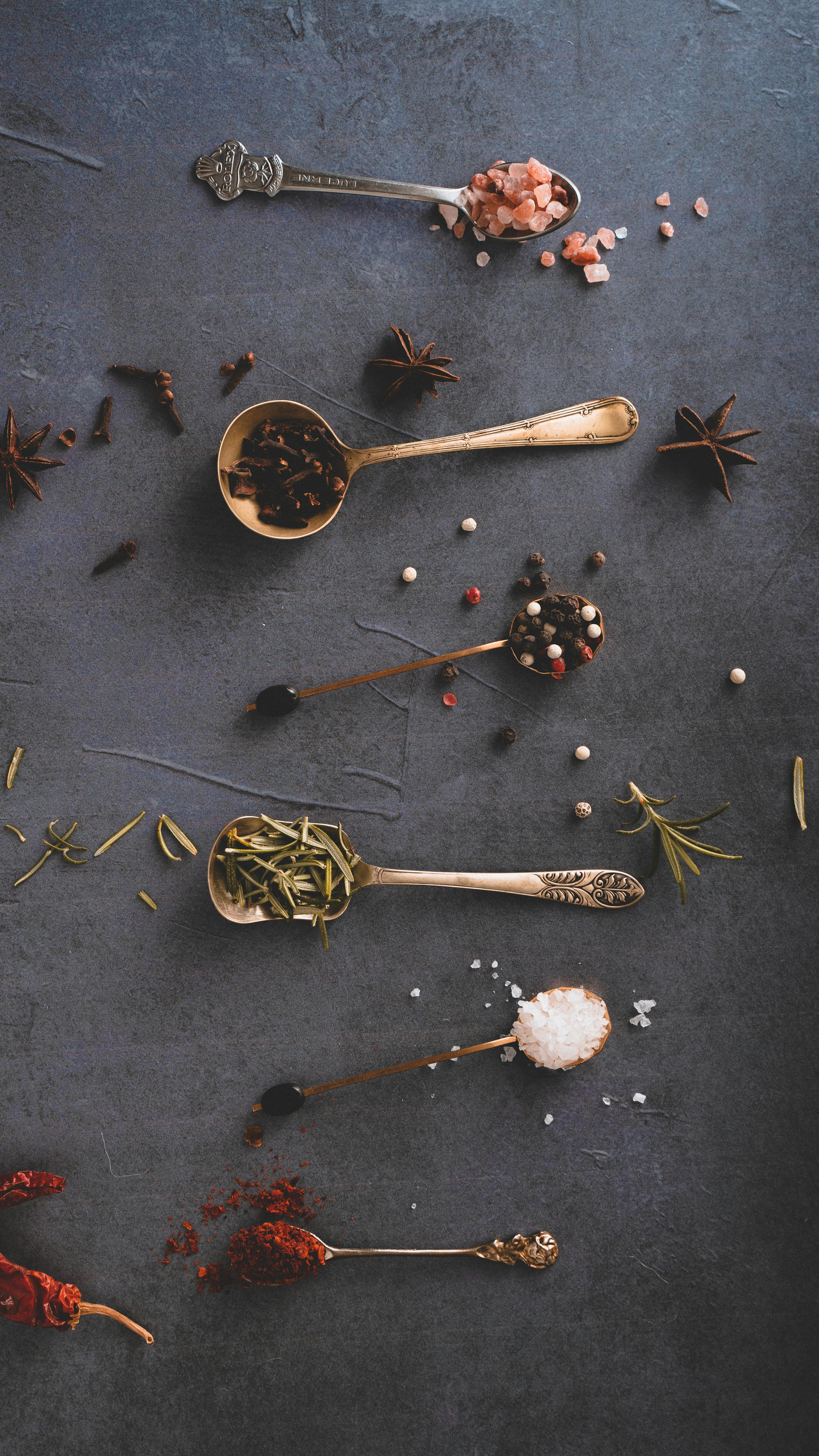 A stylish flat lay of diverse spices and herbs in golden spoons against a dark background.